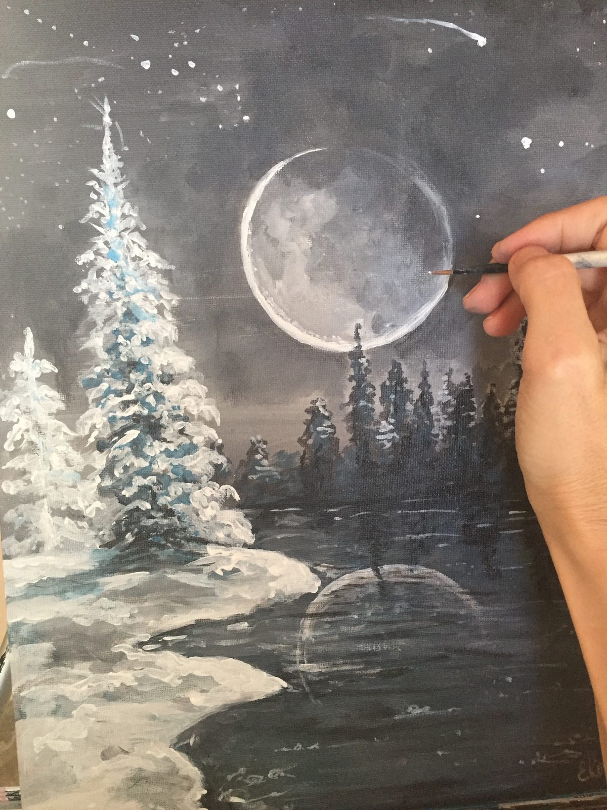 Ella's hand painting a full moon over a snowy lake at night, pine trees and the moon reflected in the dark water.