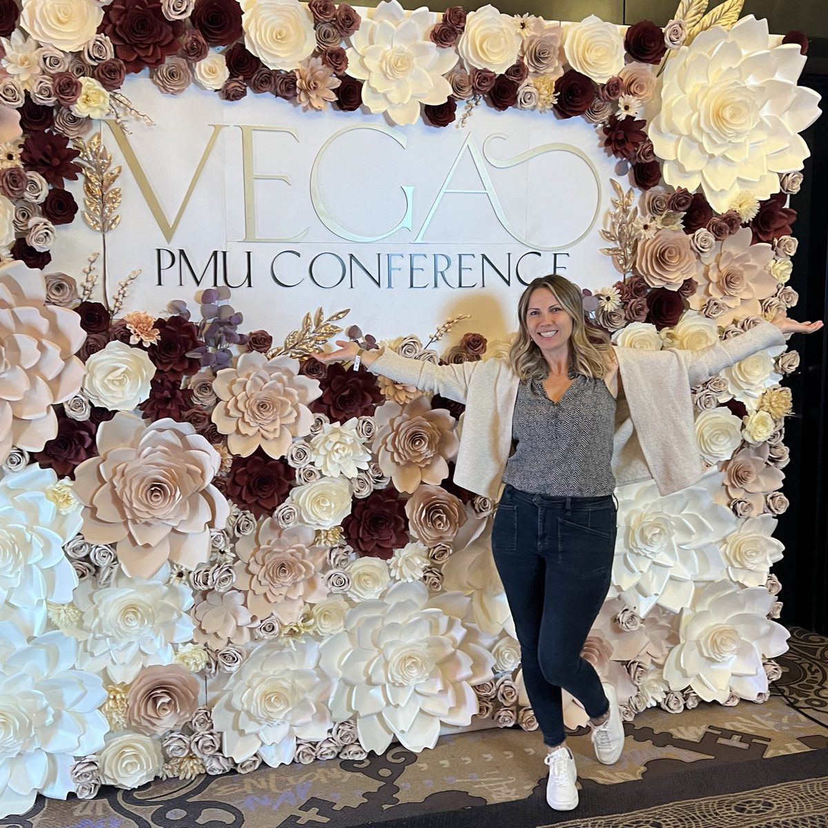 Ella, smiling with her arms out, in front of the flower wall at the Vegas PMU Conference sign in Las Vegas.