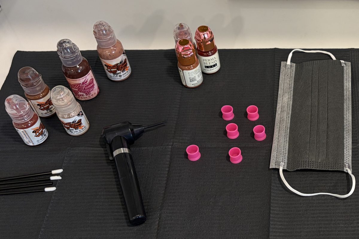 Seven pigment bottles in graduated skin tones laid out on a black drape with a shading tool, six pink pigment cups, cotton swabs and a face mask.