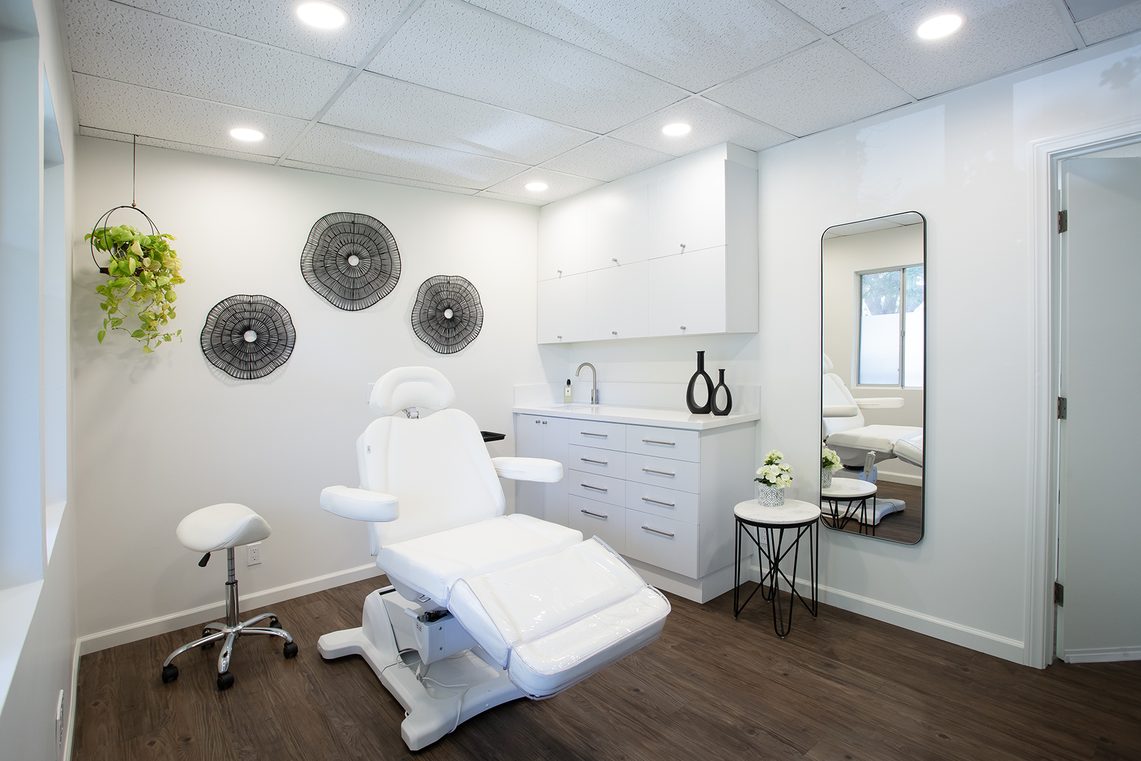The studio seen from the doorway: white reclining chair, wheeled stool, white cabinets and sink, a full-length mirror and dark wood floor.