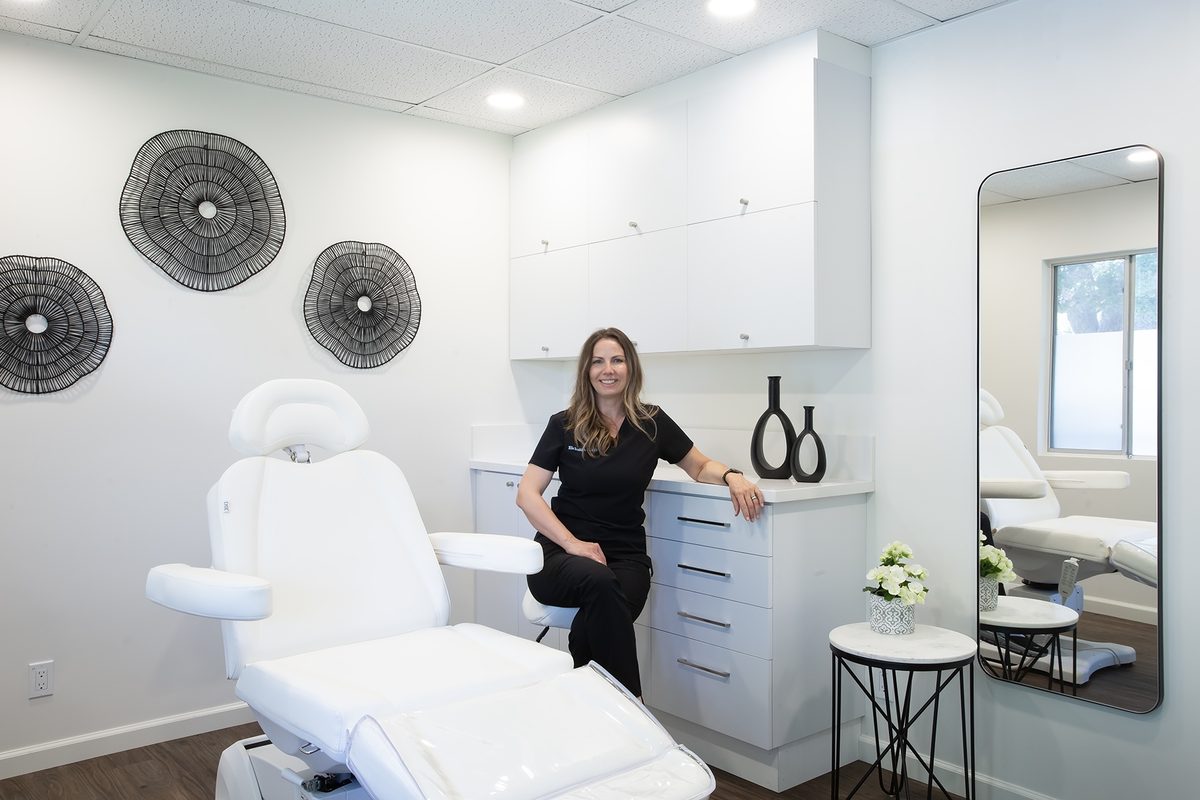 Ella sitting on the studio counter stool beside the white reclining chair, in black scrubs, in her bright white studio.